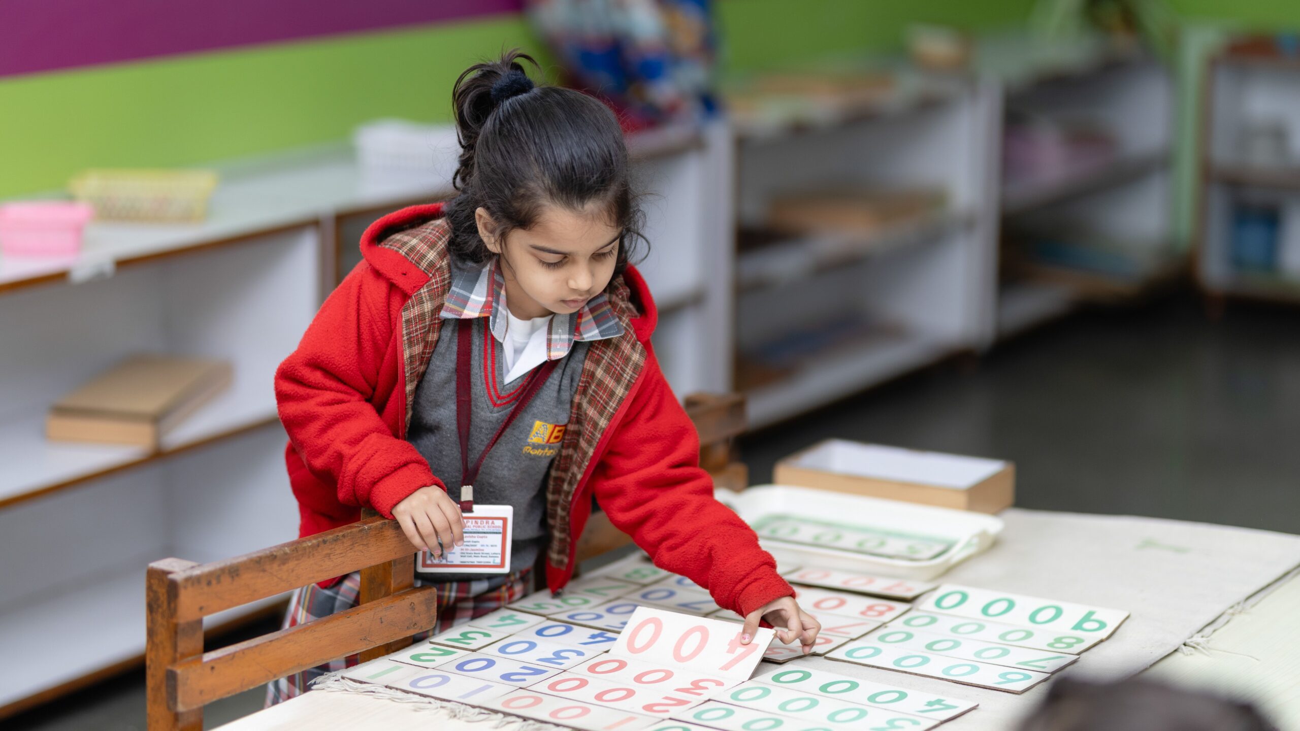 A child in a classroom wearing a red sweater looking through papers on a desk.